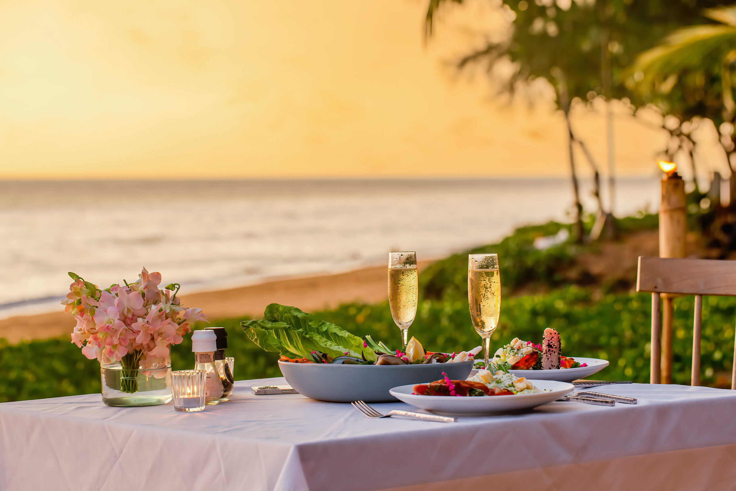 Romantic sunset dinner on beach. Table honeymoon set with luxurious food, glasses of champagne drinks in restaurant with sea view and palm trees on background. Summer vacation or wedding concept Romantic dinner by the beach at Movenpick Resort Cam Ranh's private shore.
